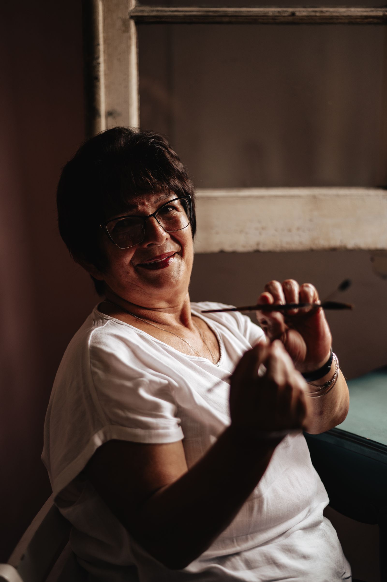 Elderly woman wearing glasses and smiling warmly with a paintbrush in hand, indoors.